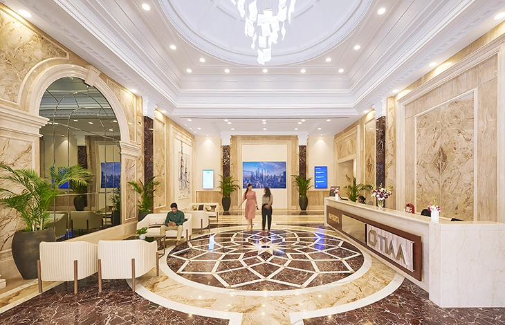 A modern office lobby with a sleek, white marble reception desk, plants on either side, and a minimalist wooden wall design. A receptionist is visible behind the desk, while a person walks past the entrance.
