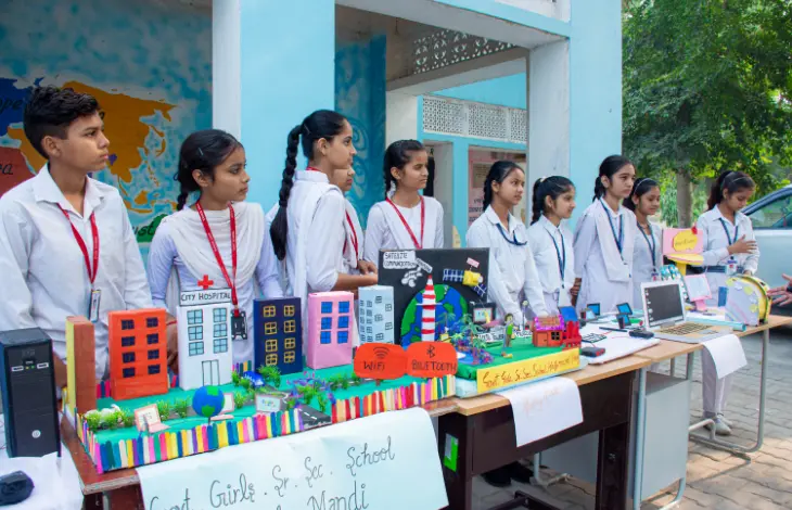 A group of school students standing beside their project display, showcasing models of buildings and technology, at a school exhibition.
