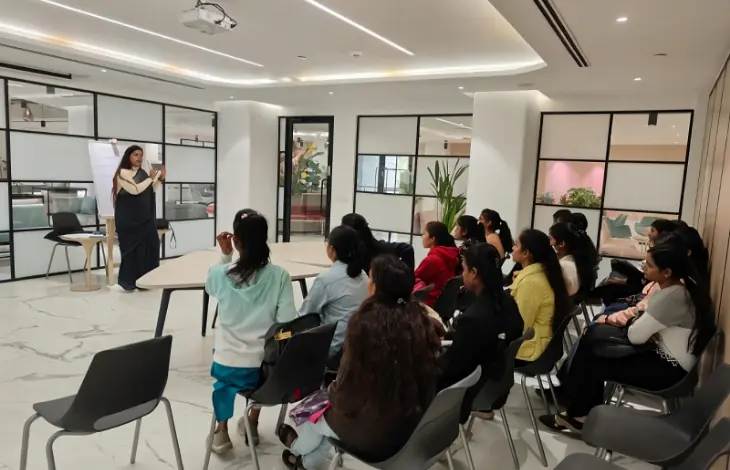 A woman leading a session with a group of people seated and listening attentively in a modern office setting.
