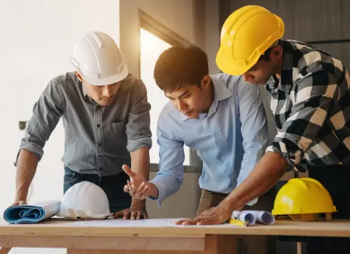 Three construction professionals in safety helmets reviewing blueprints and discussing plans around a wooden table.