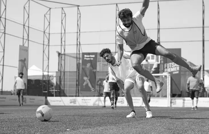 A black-and-white image of two players during a football match, one of them in mid-air after a fall while attempting to reach the ball.
