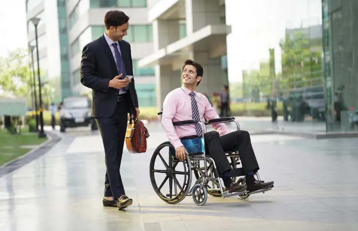Two professionals, one in a wheelchair and the other walking beside him, smiling and having a conversation outside a modern office building