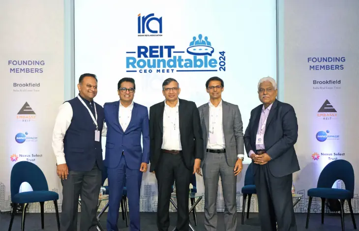 A group of five professionals standing together for a photo at the REIT Roundtable CEO Meet 2024, with the event backdrop featuring the logos of participating members