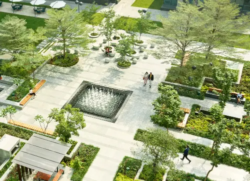 A high-angle view of a vibrant landscaped area with a central water fountain, surrounded by well-maintained greenery, pathways, and seating spaces. People are walking through the space, enjoying the peaceful environment