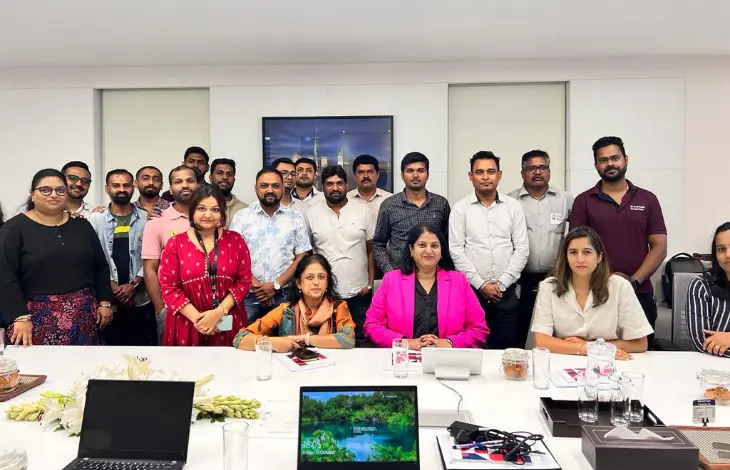A group photo of employees in a meeting room, with a mix of men and women, some seated and others standing, around a conference table