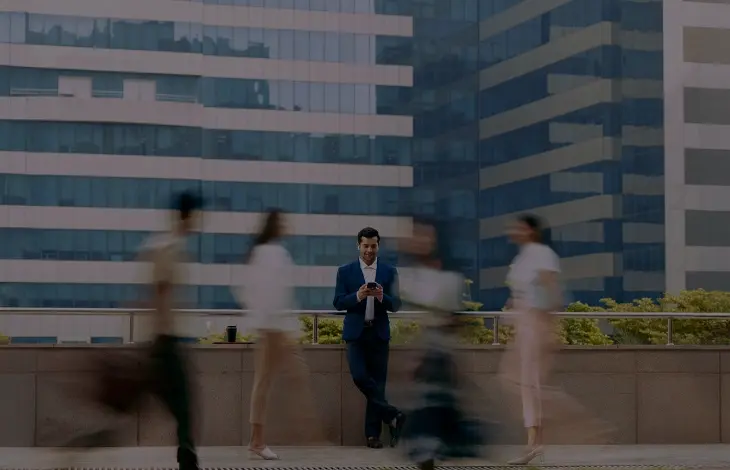 A professional man in a suit using his phone, standing still amid blurred motion of passersby, with modern office buildings in the background at Candor TechSpace, Sector 135, Noida.