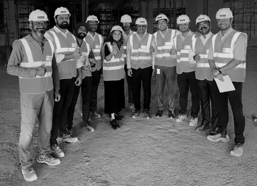 A group of 12 construction workers in high-visibility vests and hard hats, posing together on a construction site. They are smiling and giving thumbs up, with the background showing an unfinished building structure.