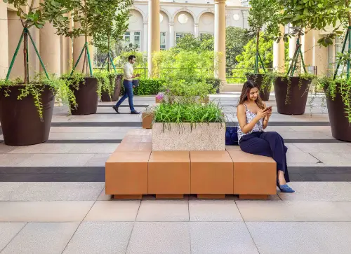 A woman sitting on a bench at Spectra Podium in Downtown Powai, Mumbai, while texting, with a man walking by in the background. Large potted plants add greenery to the urban space