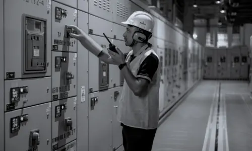 Black and white image of a technician wearing a hard hat and safety vest, operating electrical control panels in an industrial setting.