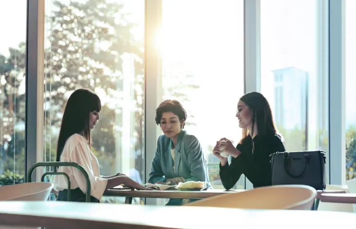 Three women having a discussion over coffee in a bright, modern office setting, with sunlight streaming through large windows.